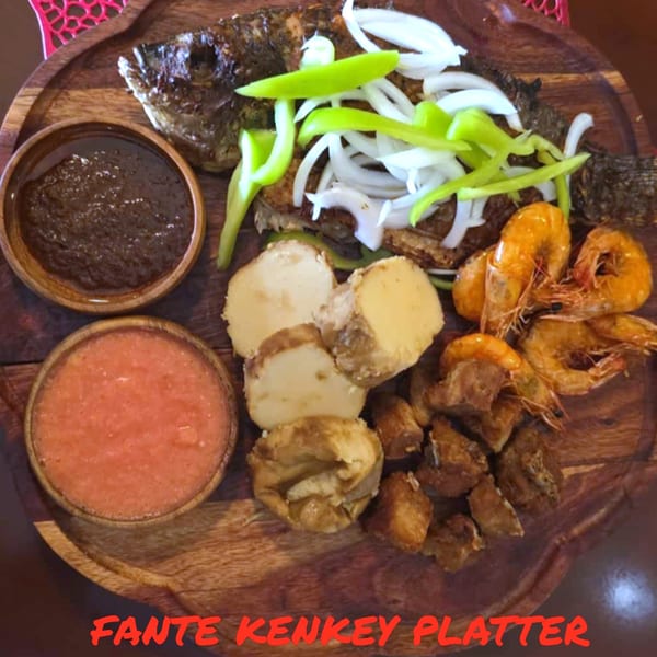 A Fante Kenkey Platter with grilled fish, shrimp, kenkey, and two sauces on a wooden board.