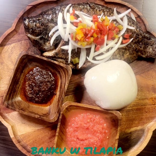 Grilled tilapia with sliced onions, bell peppers, banku, and two sauces on a wooden platter.